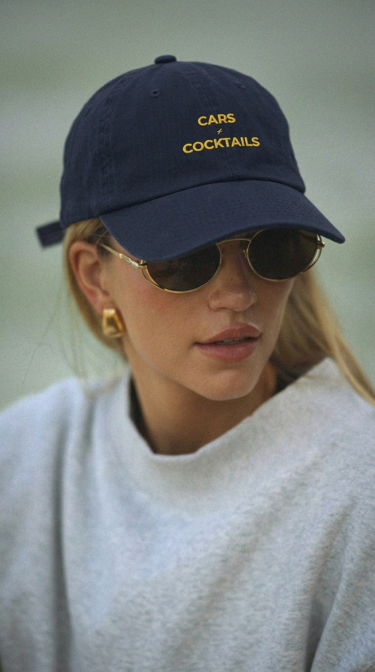 A woman wearing a navy blue baseball cap with yellow embroidered text reading 'CARS + COCKTAILS', along with round sunglasses and a light gray sweatshirt, photographed outdoors against a soft blurred background.